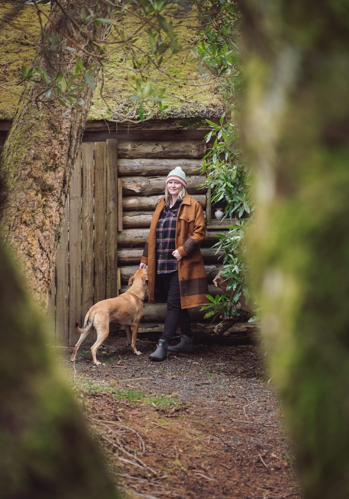 Scottish storyteller Claire Gibb of Lullabrae photographed outdoors with her dog beside a rustic woodland cabin, framed by mossy trees and natural greenery. Branding photograph by Ayrshire photographer Renata Clarke.